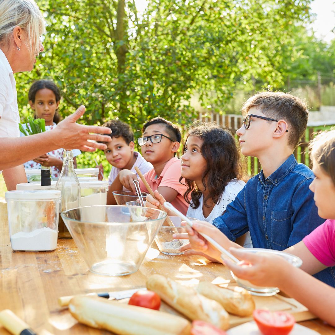 Kids and a teacher outside being instructed on how to make a dish 