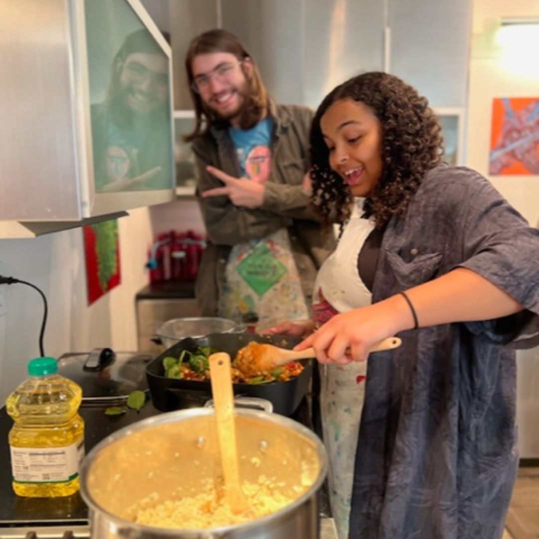 two people are in this image. one is cooking on the stove top in the foreground. in the background  to the side is another person giving a peace sign. The two are apart of the cooking class being described in this article. 